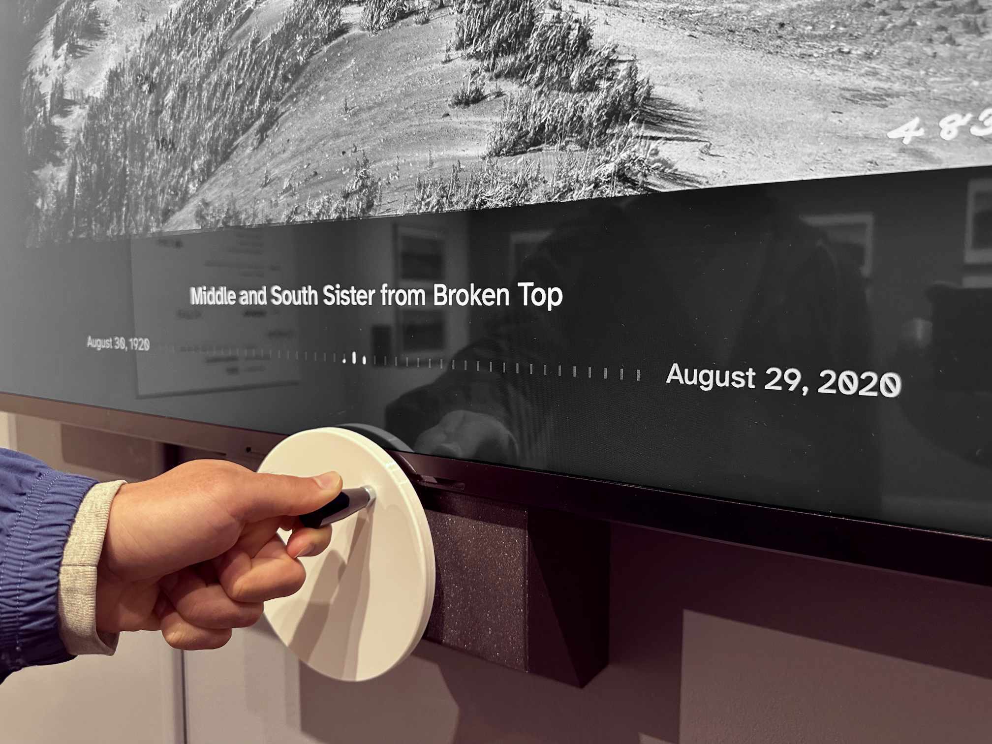 A visitor’s hand turning the pale wooden crank beneath a panoramic screen captioned “Middle and South Sister from Broken Top, August 29, 2020”.