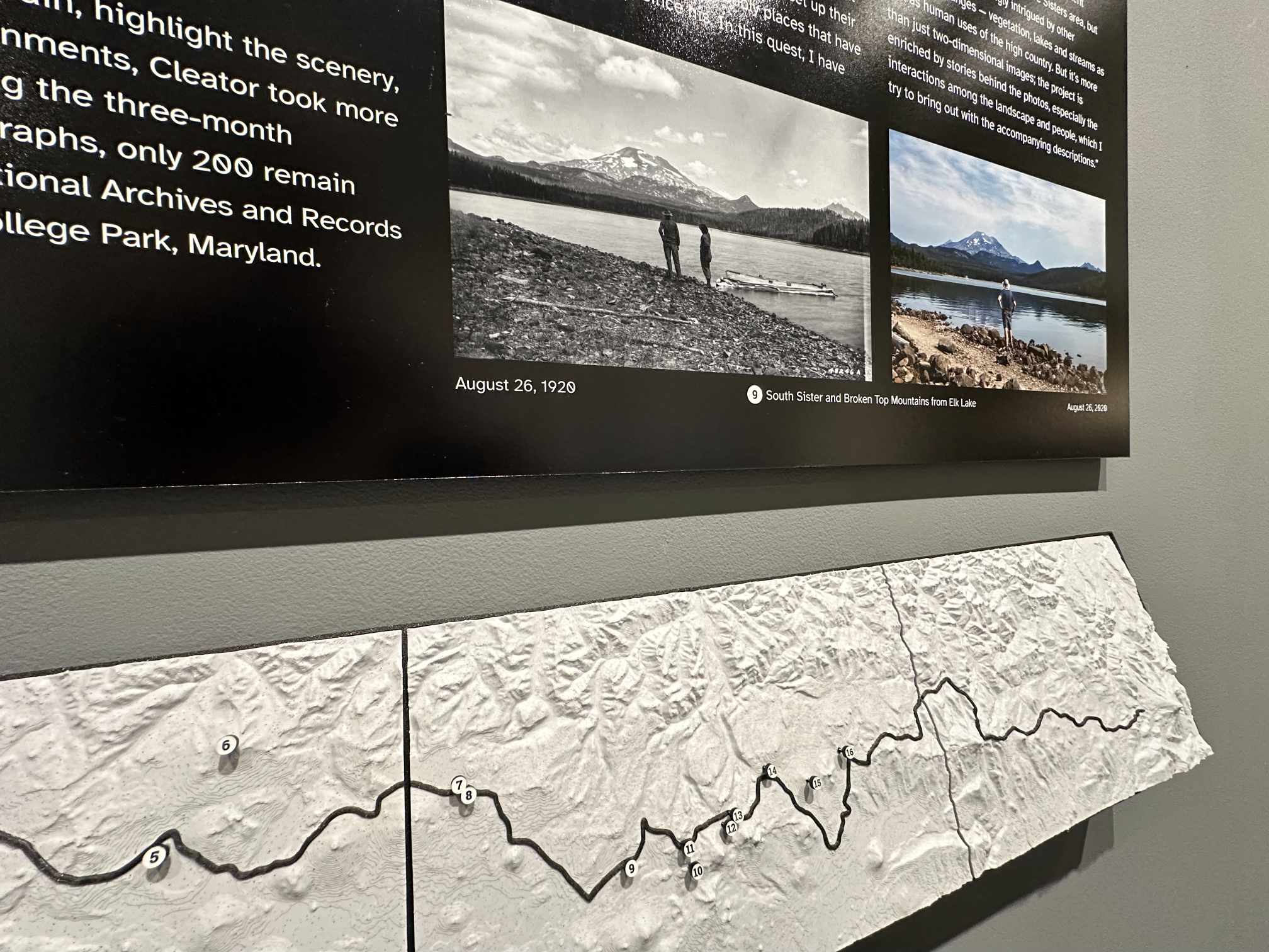The tactile topographic map, a raised relief panel with the trail routed across it as a black line, set below wall text and paired photographs.